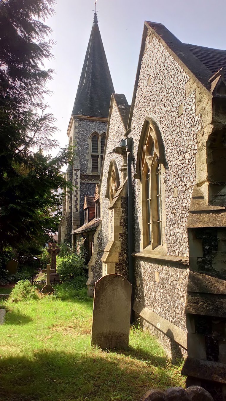 Golden evening light on the flint walls and spire of St Nicholas Church, with churchyard graves